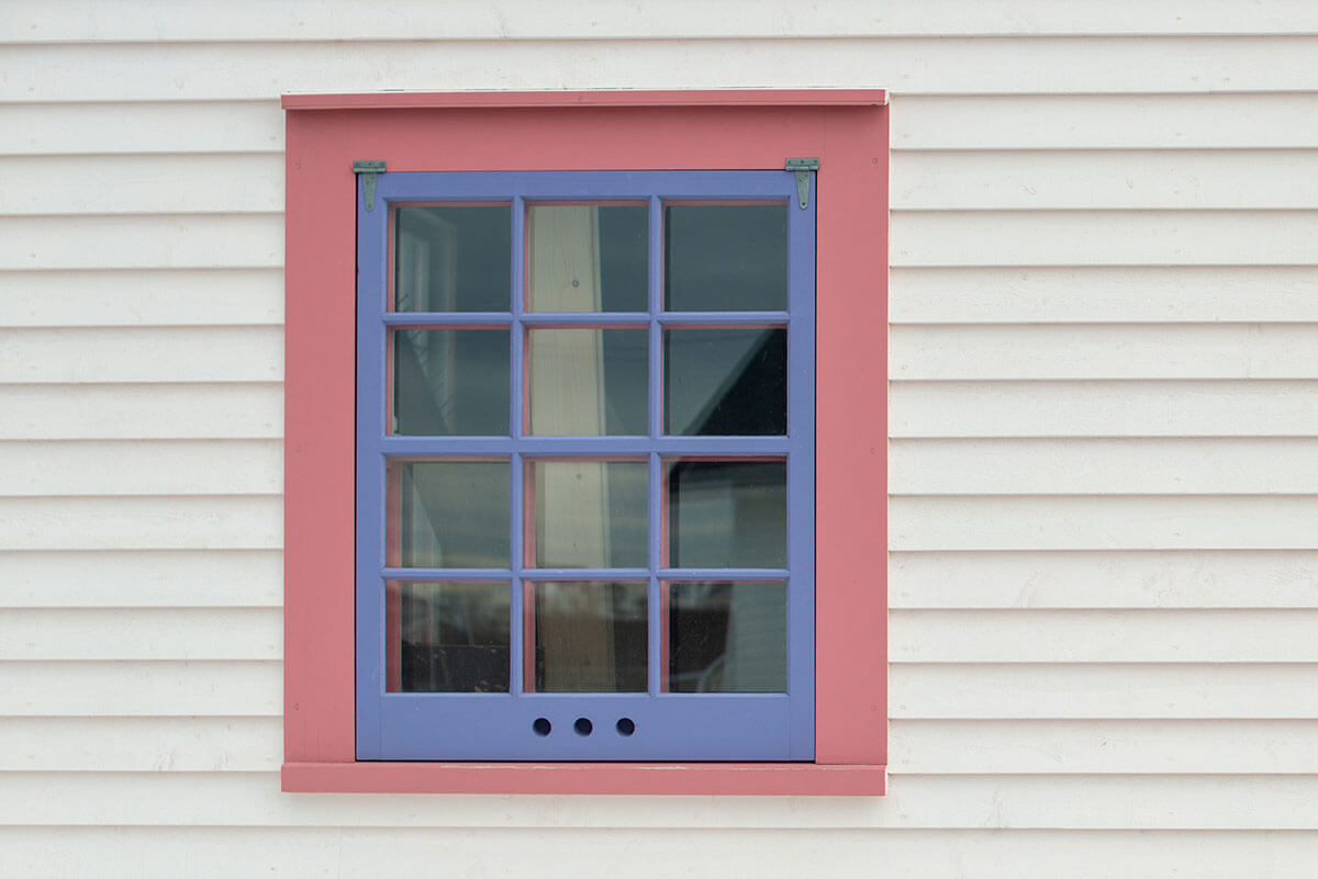 A single vintage storm window with 12 small panes, a purple wooden frame, and pink trim in a white wooden clapboard building. The exterior wall is made of narrow pine horizontal clapboard siding.